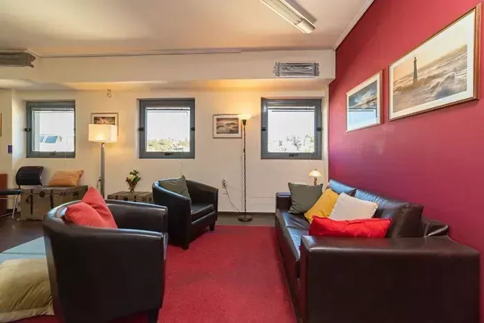 Ένα σαλόνι με δερμάτινα έπιπλα, κόκκινο χαλί και τοίχους με κόκκινες πινελιές διακοσμημένους με πλαισιωμένες εικόνες τοπίων.