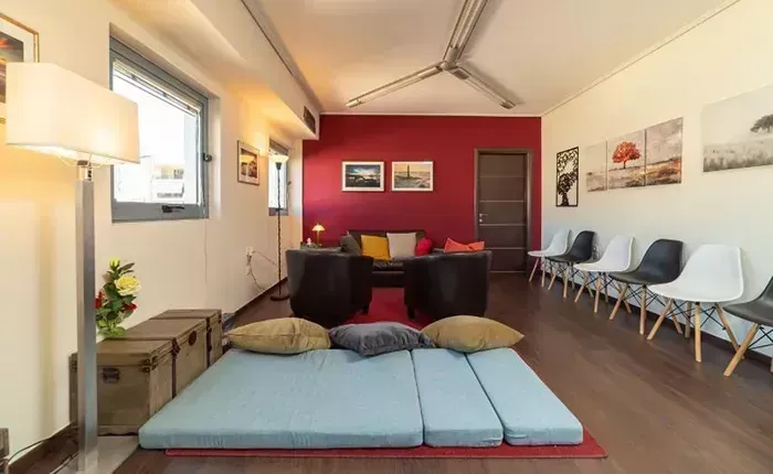 Μια μοντέρνα αίθουσα αναμονής διαθέτει ένα ανοιχτό μπλε στρώμα δαπέδου με μαξιλάρια, σκούρες πολυθρόνες και μια σειρά από καρέκλες κατά μήκος ενός τοίχου.