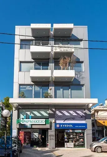 Ένα πολυώροφο γκρι κτίριο με στοιβαγμένα λευκά μπαλκόνια, που διαθέτει ένα φαρμακείο και ένα κατάστημα γυαλιών στο ισόγειο.