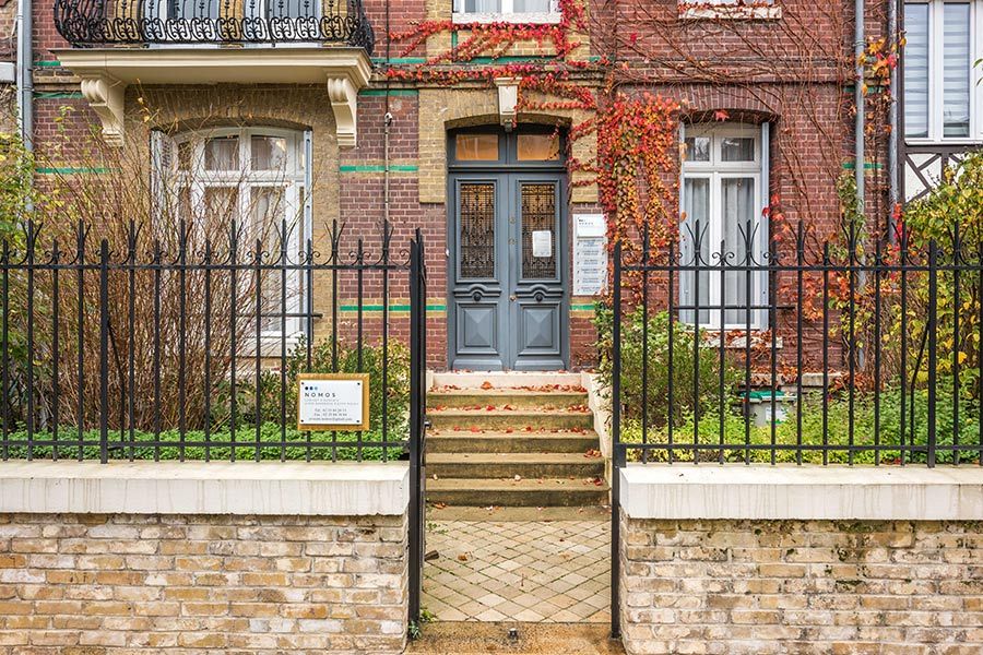 Bâtiment en briques avec clôture en fer forgé, porte grise et marches menant à l'entrée.