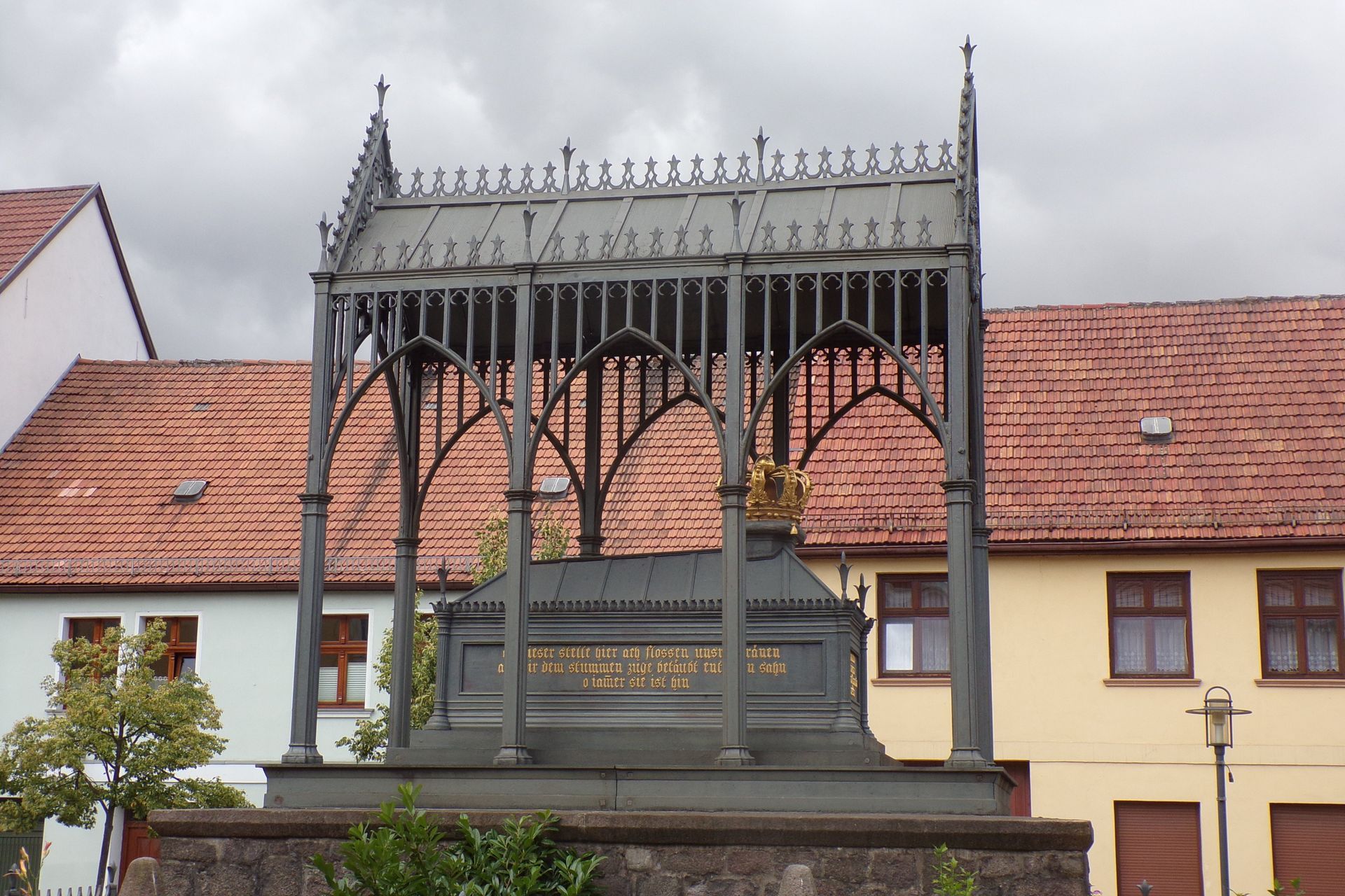 Grabdenkmal mit kunstvollem schmiedeeisernen Baldachin vor orangefarbenen Gebäuden unter bewölktem Himmel