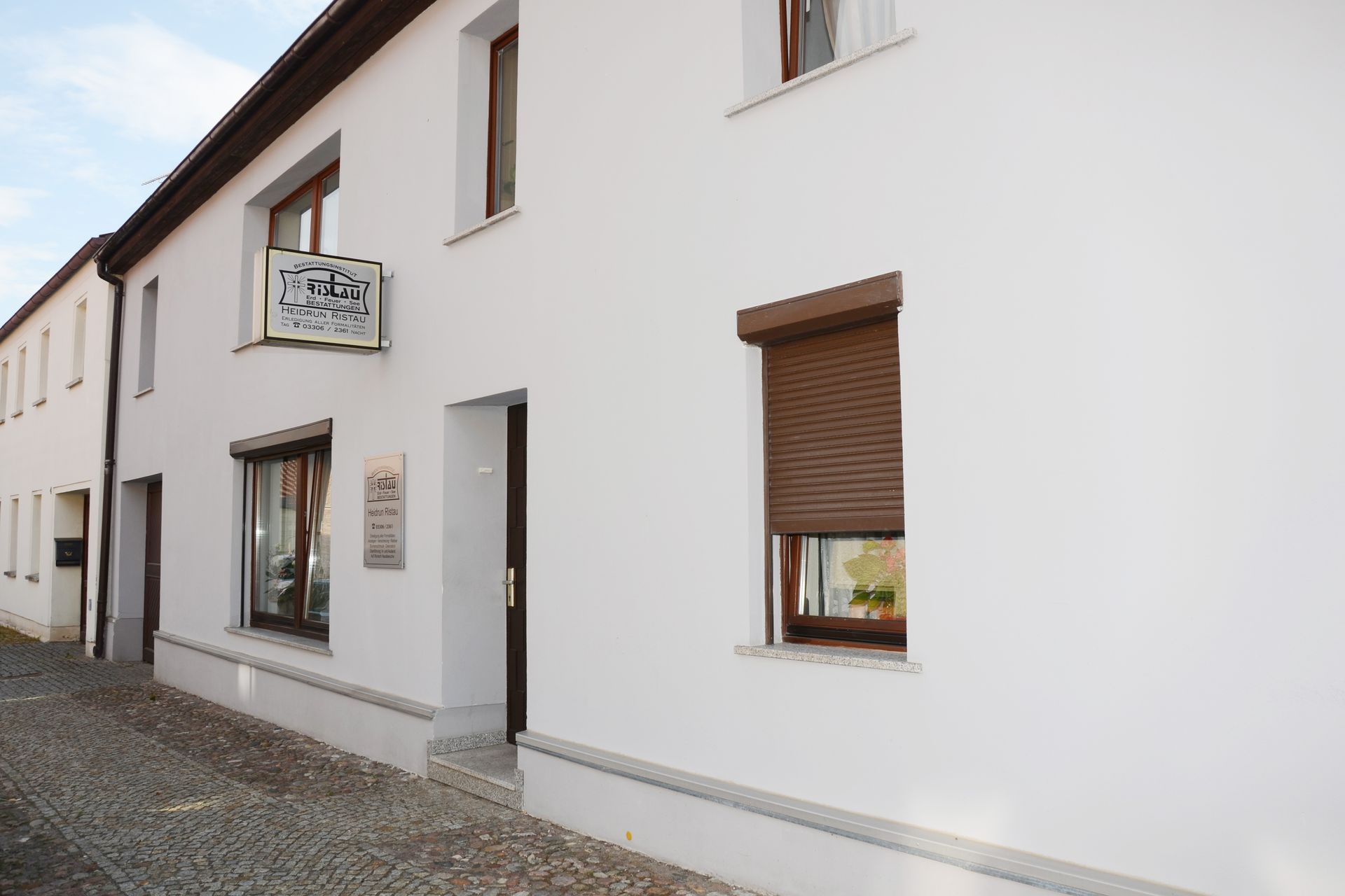 Weißes Gebäude mit kleinem Schild, braunen Fensterläden und gepflastertem Gehweg
