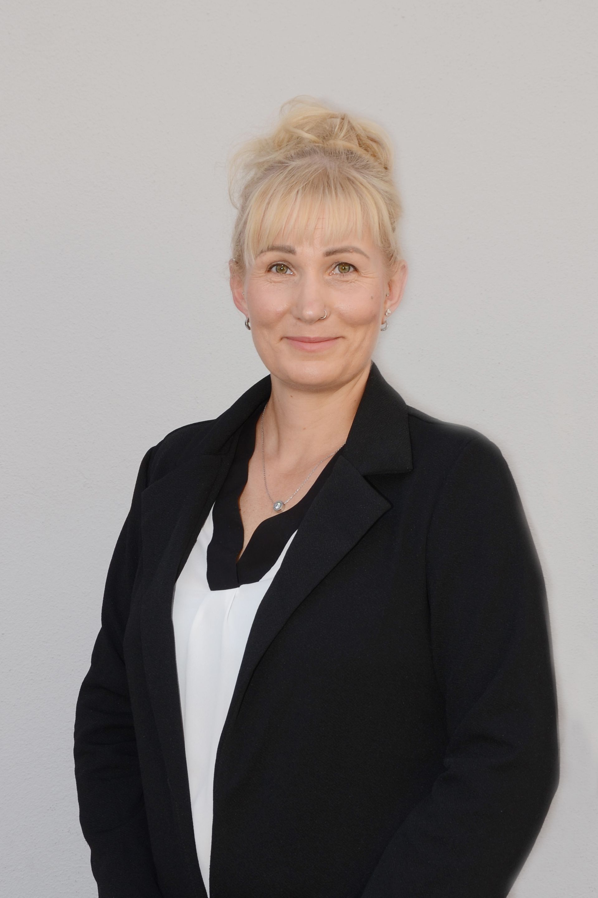 Frau mit blonden Haaren in schwarzem Blazer und weißer Bluse vor grauem Hintergrund, lächelnd.