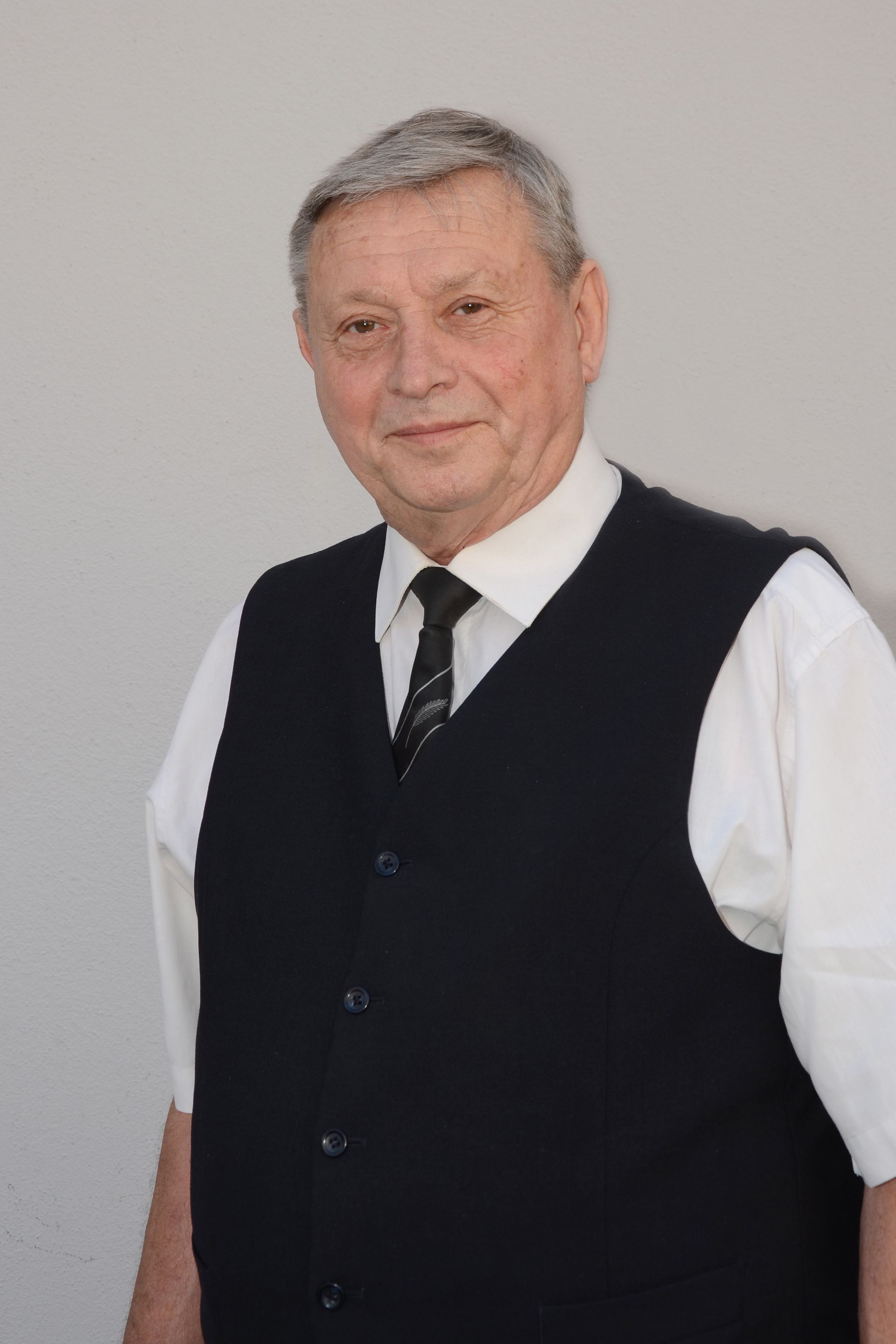 Älterer Mann in Weste und Krawatte, lächelnd vor einer weißen Wand.