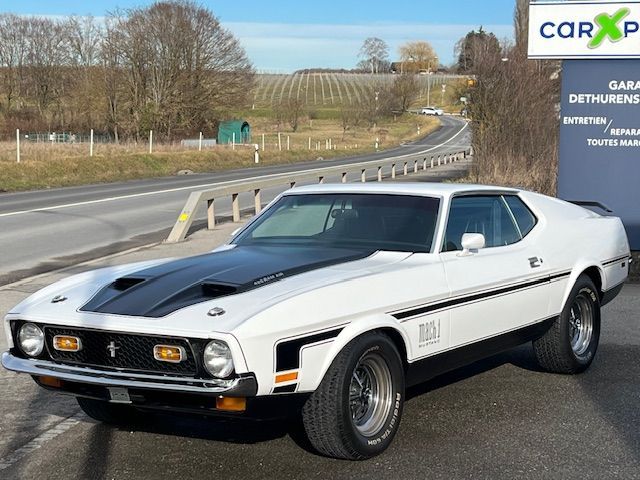 Ford Mustang Mach 1 de 1971, blanche et noire, garée au bord d'une route, avec un capot et des bandes latérales noires.