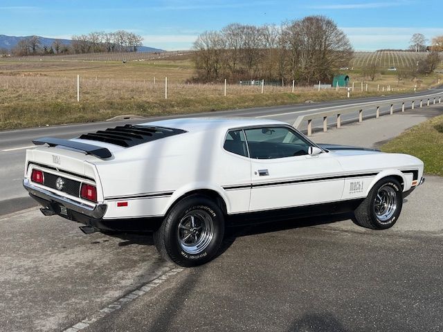 Ford Mustang Mach 1 blanche de 1971, muscle car, sur une route, bandes noires, persiennes sur la lunette arrière.