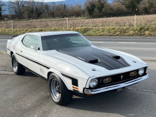 Ford Mustang Mach 1 de 1971, blanche et noire, garée sur le trottoir. Capot et bande de course noirs.