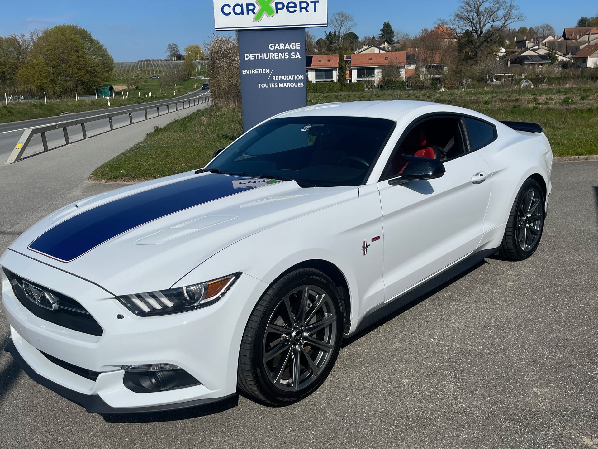 Une Ford Mustang blanche à bandes de course bleues est garée à l'extérieur, près d'un panneau de concessionnaire.