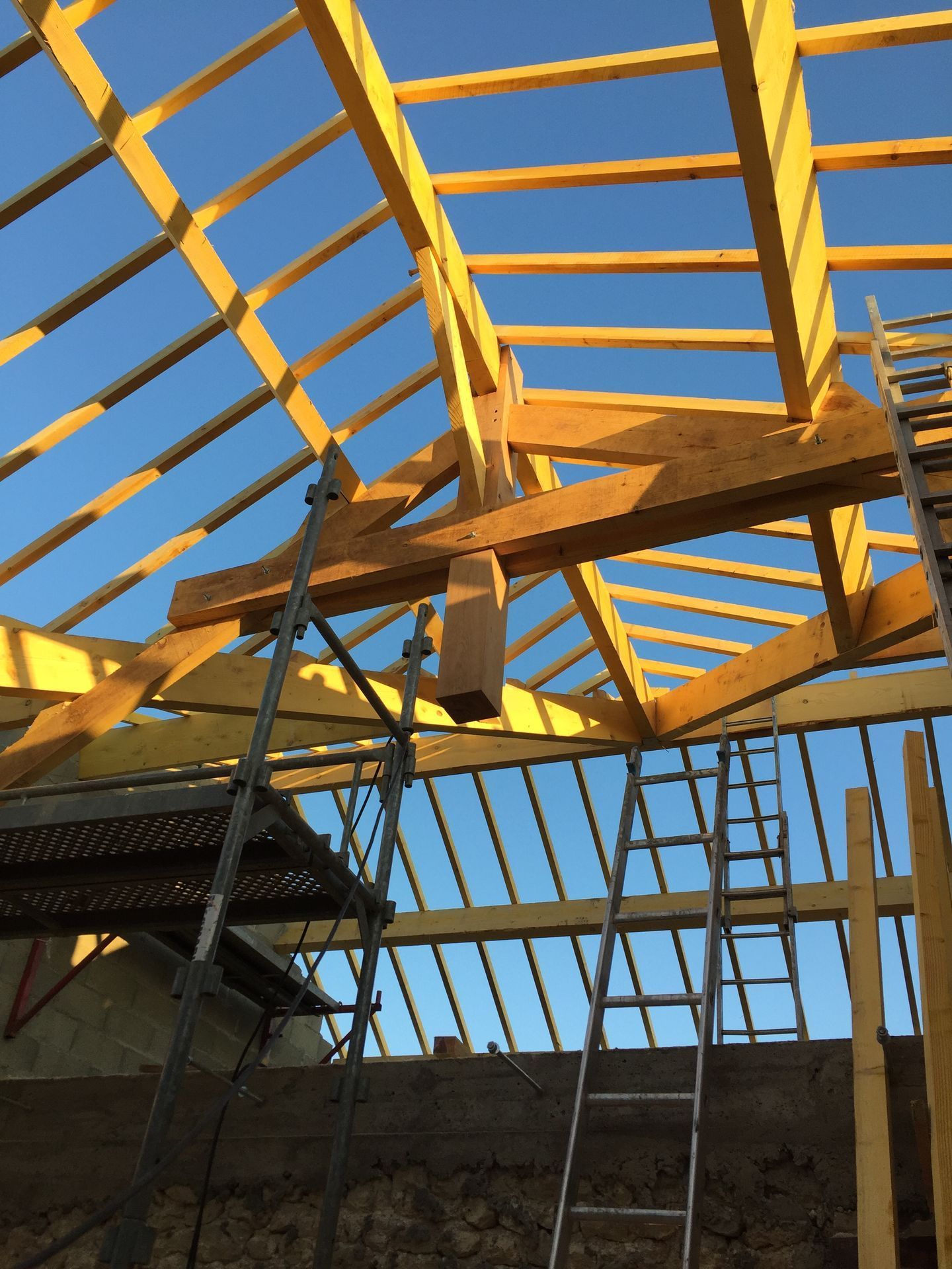 Charpente de toit en bois en construction sur fond de ciel bleu, avec une échelle en dessous.