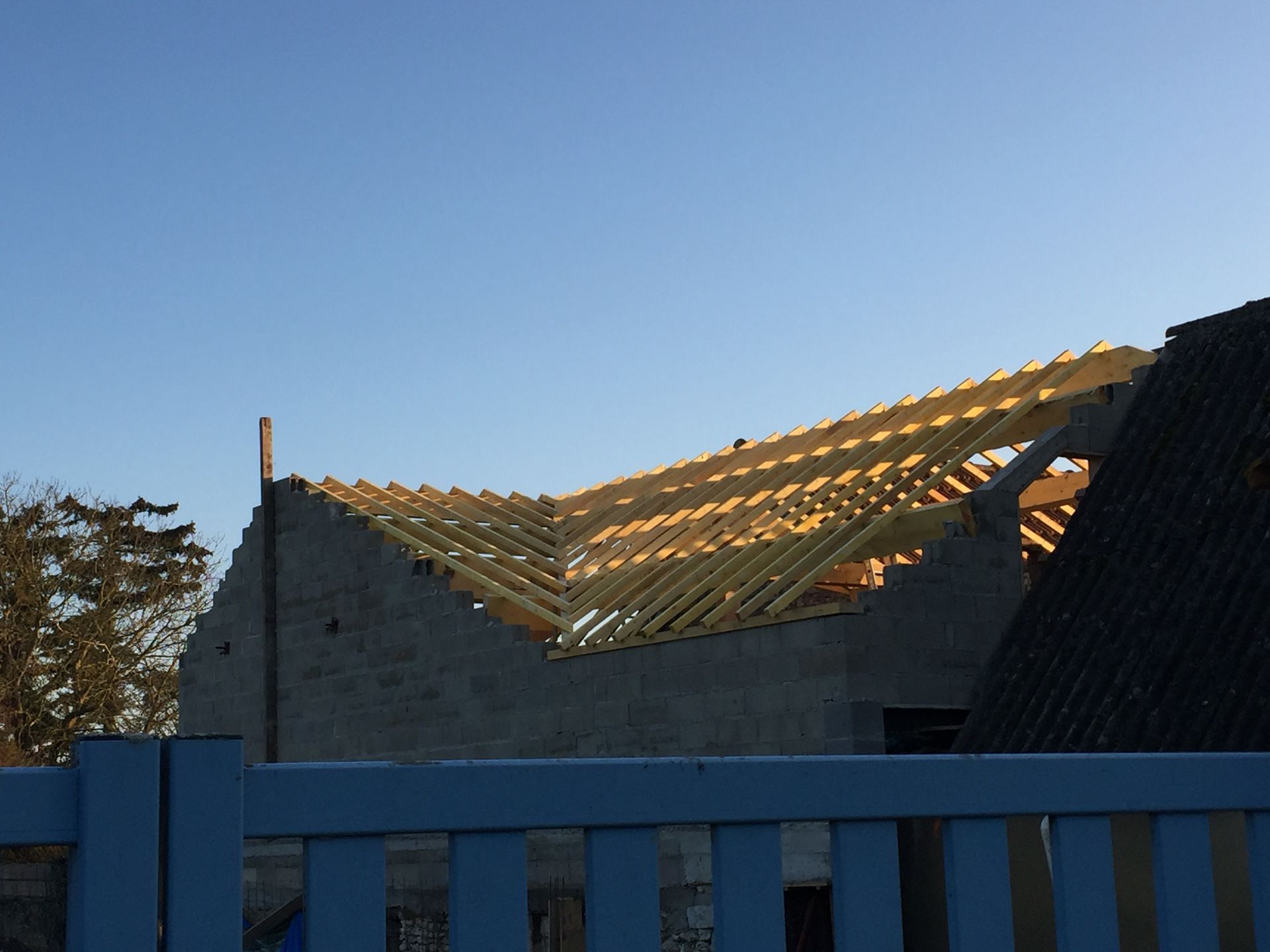 Toiture partiellement construite avec poutres en bois apparentes au-dessus d'une clôture bleue au coucher du soleil