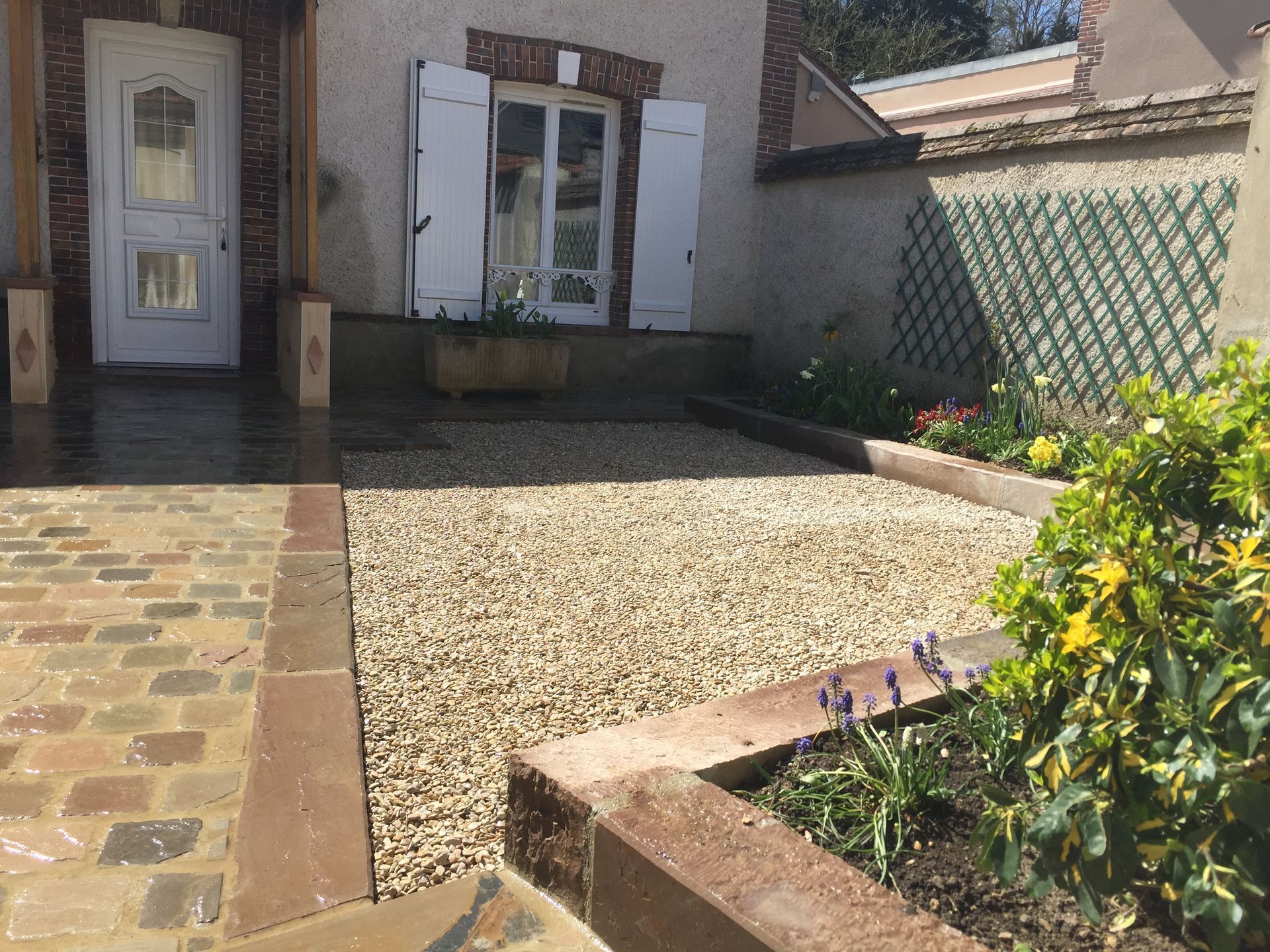 Cour d'entrée principale avec volets blancs ouverts, chemin pavé et petite terrasse en gravier bordée de verdure.