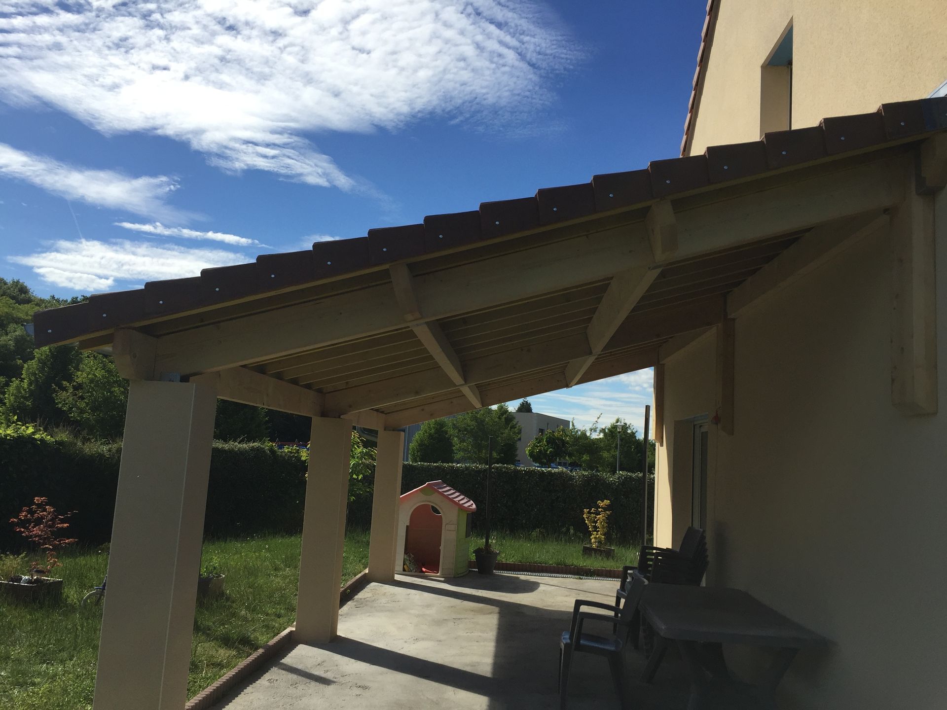 Terrasse couverte attenante à une maison, avec chaises, table et jardin gazonné sous un ciel bleu.