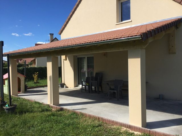 Terrasse couverte d'une maison beige avec colonnes, chaises et jardin gazonné sous un ciel bleu.