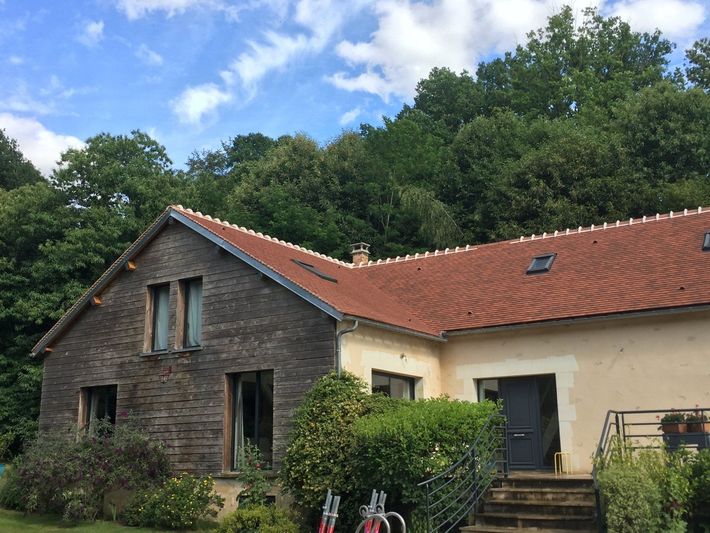Maison en briques au toit de tuiles rouges et aux haies vertes, se détachant sur un fond d'arbres sous un ciel partiellement nuageux.