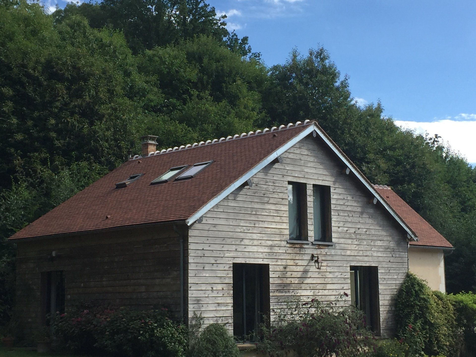 Petite maison en pierre au toit de tuiles rouges et aux puits de lumière, située au milieu d'une dense végétation.