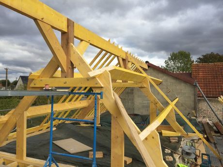 Charpente de toit en bois jaune en construction sur une maison, sous un ciel nuageux, avec des échafaudages à proximité.