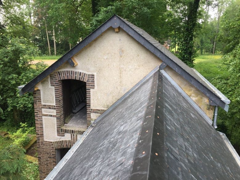 Petit bâtiment en briques et plâtre avec un toit gris pentu dans une zone boisée et feuillue