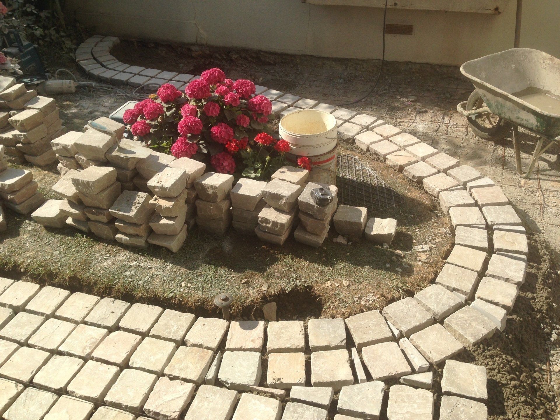 Bordure de jardin en pierre incurvée entourant un petit parterre de fleurs roses et des pavés empilés à proximité
