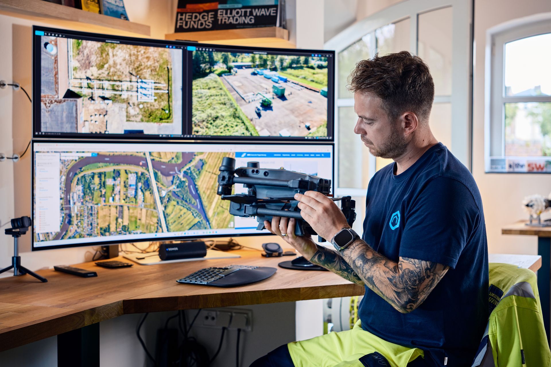 Man holding drone, looking at it, with monitors displaying aerial views, sitting at a desk.