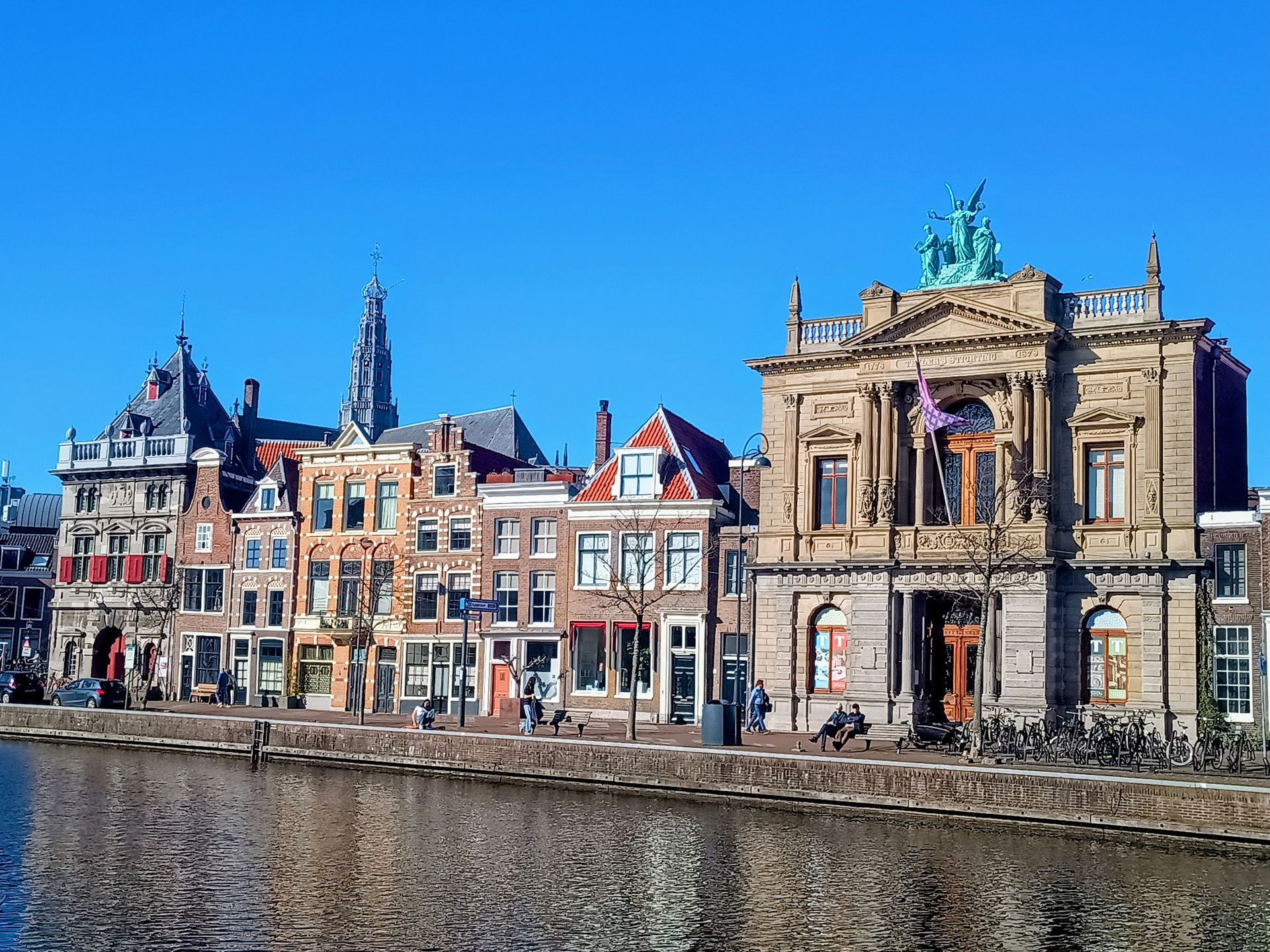 Historic canal in Haarlem with colorful gabled houses, a grand classical building, bicycles along the quay, and reflections shimmering in the calm water.