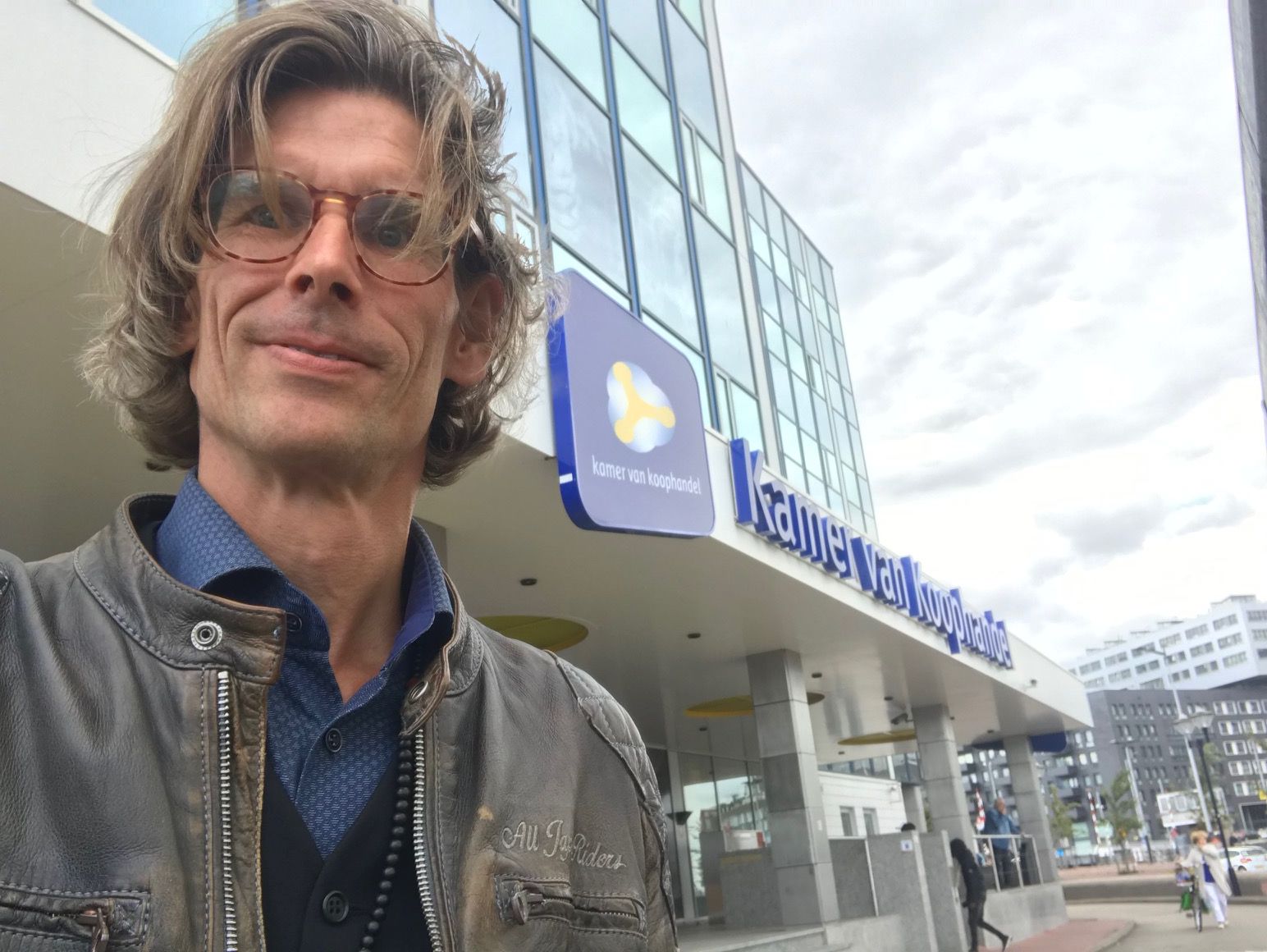 Sander Faass wearing glasses and a leather jacket standing outside the Chamber of Commerce (Kamer van Koophandel) building in the Netherlands.