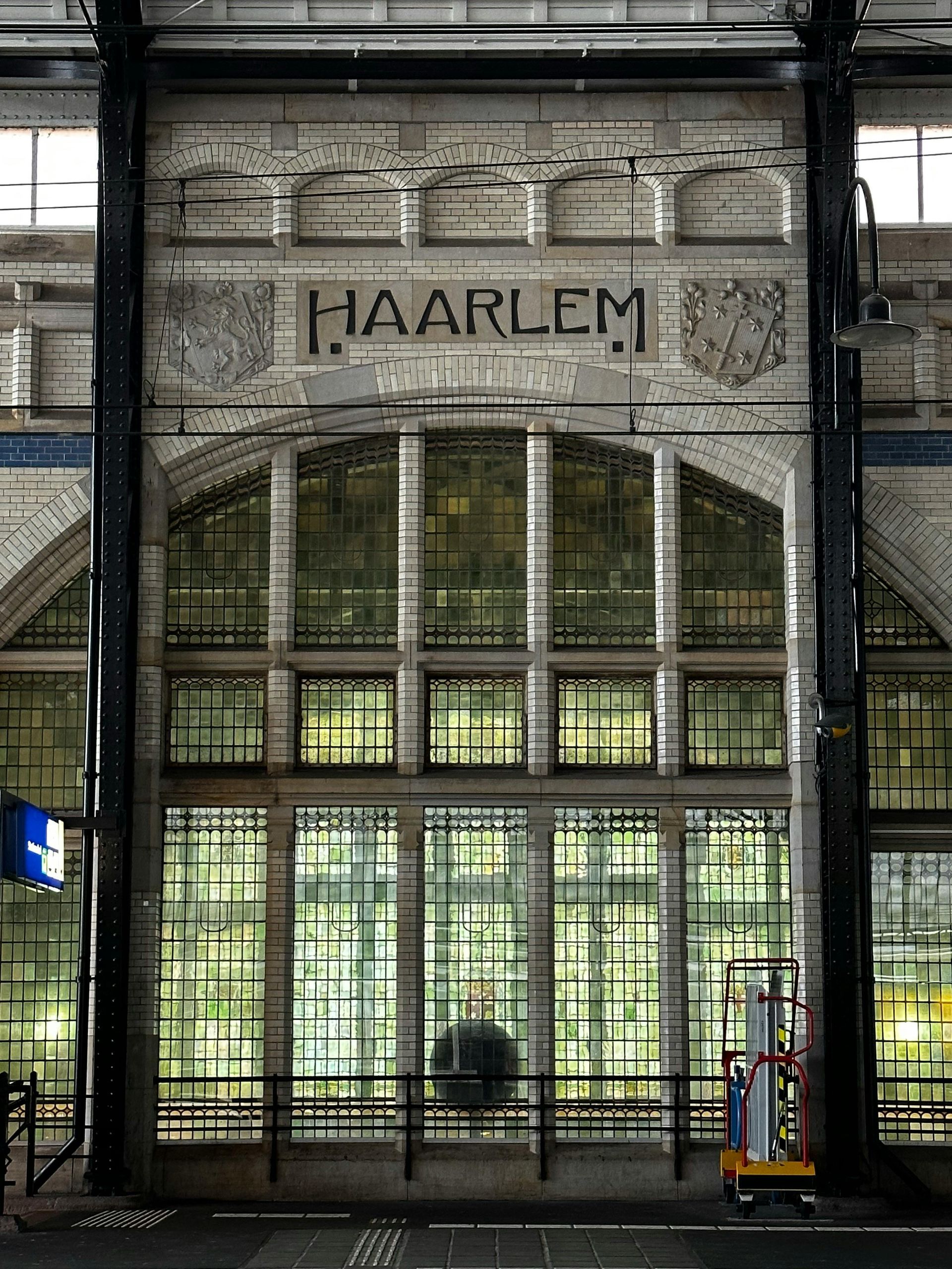 Historic train station hall with a tall brick façade and large arched stained-glass windows. The word “HAARLEM” appears above the windows, flanked by two coats of arms. Dark steel beams and overhead railway lines run in front of the building.