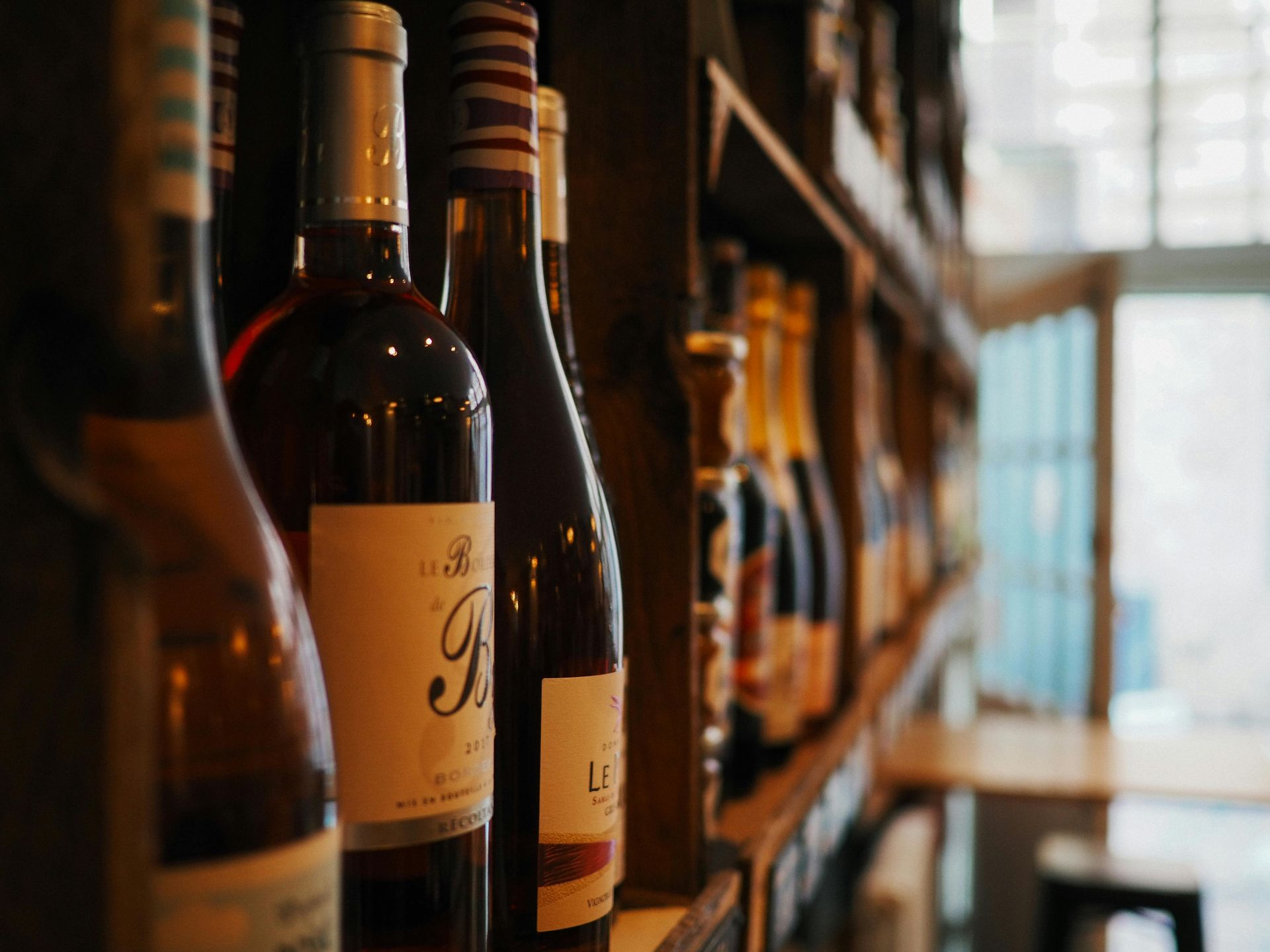 Close-up of wine bottles displayed on a wooden shelf, with warm ambient lighting highlighting the labels and glass reflections in a cozy interior setting.