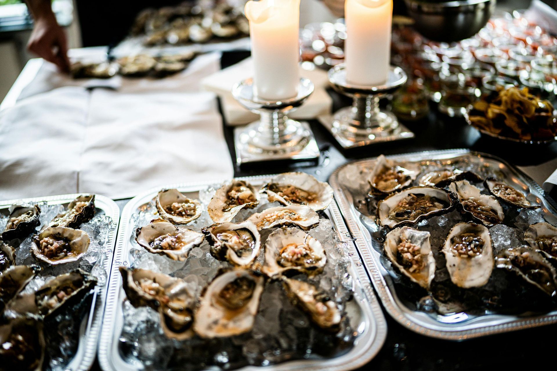 Platters of fresh oysters served on crushed ice, topped with garnish, arranged on a table with lit candles and small dishes in the background, creating an elegant dining atmosphere.
