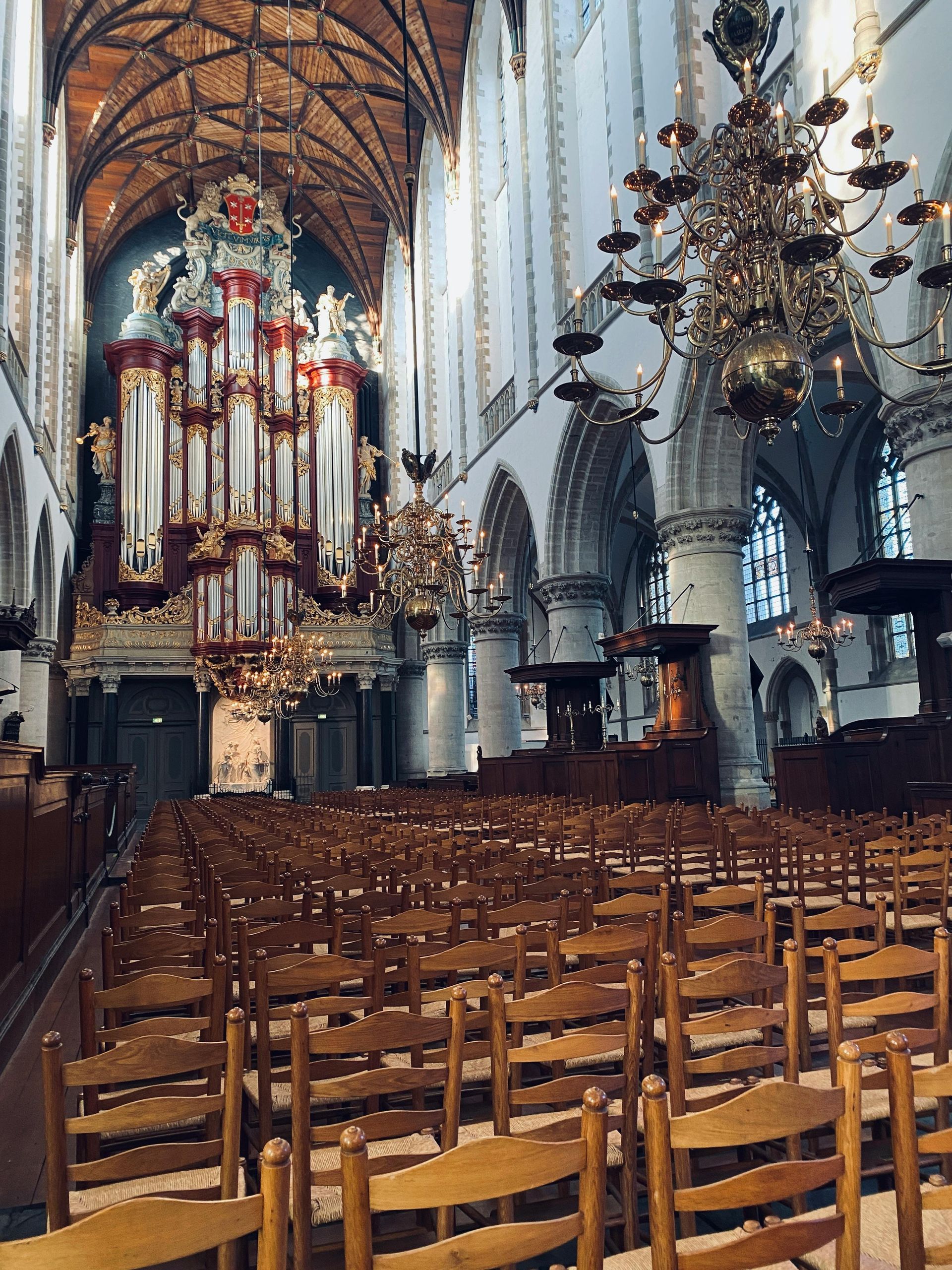 Interior of a historic church with high vaulted ceilings, large stone columns, ornate chandeliers, and rows of wooden chairs. A grand pipe organ with red and gold detailing stands prominently at the front beneath tall arched windows. St. Bavo Church Haarlem
