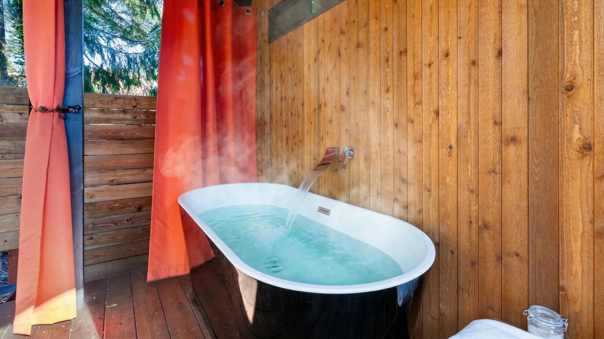 Open-air wooden bath with a white tub, orange curtains, and water inside