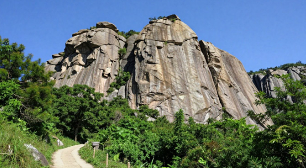 Rocky cliff face above a dirt path, surrounded by green trees under a blue sky