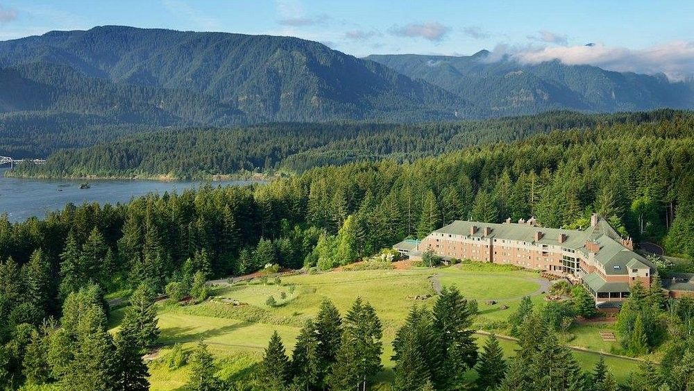 A sprawling lodge building sits on a lush green lawn bordered by a forest and a large lake, with mountains in the distance.