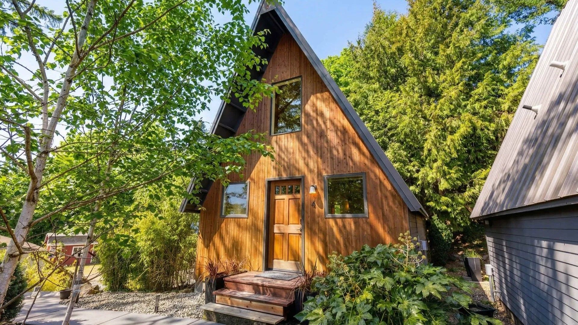 A-frame wooden cabin with a steep roof, surrounded by trees and greenery on a sunny day.