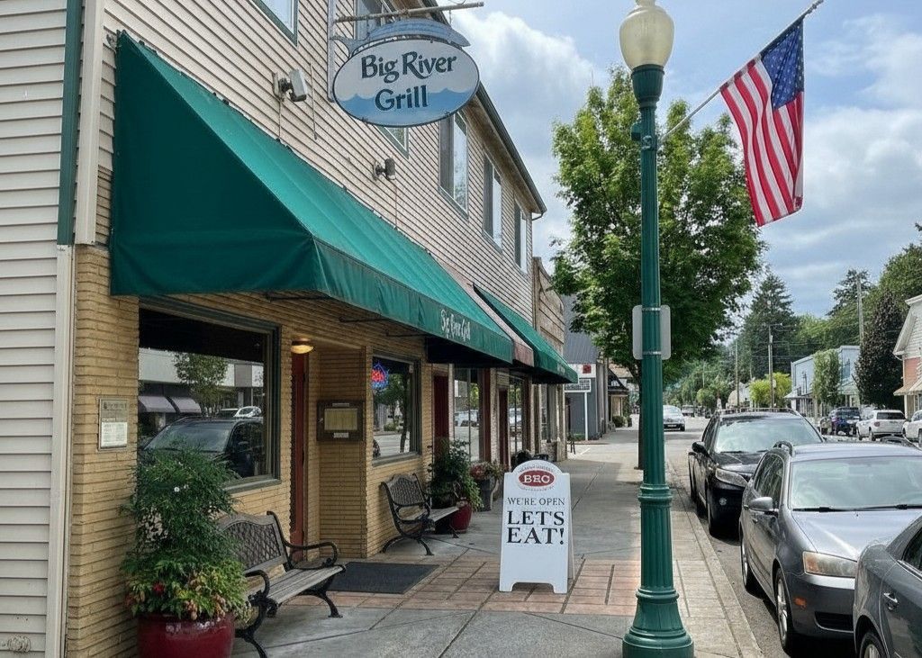 The Big River Grill storefront features a green awning, a white sidewalk sign, and an American flag on a streetlamp.