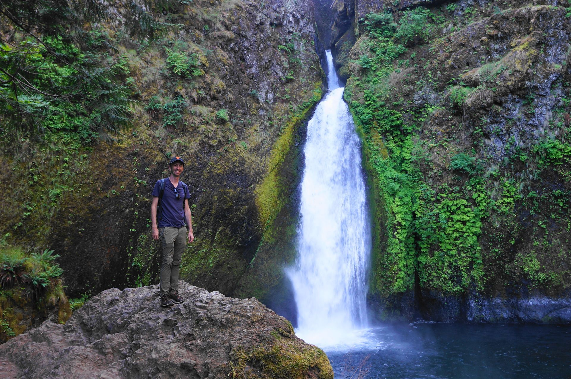 Person standing on a rock beside a waterfall in a green forest canyon