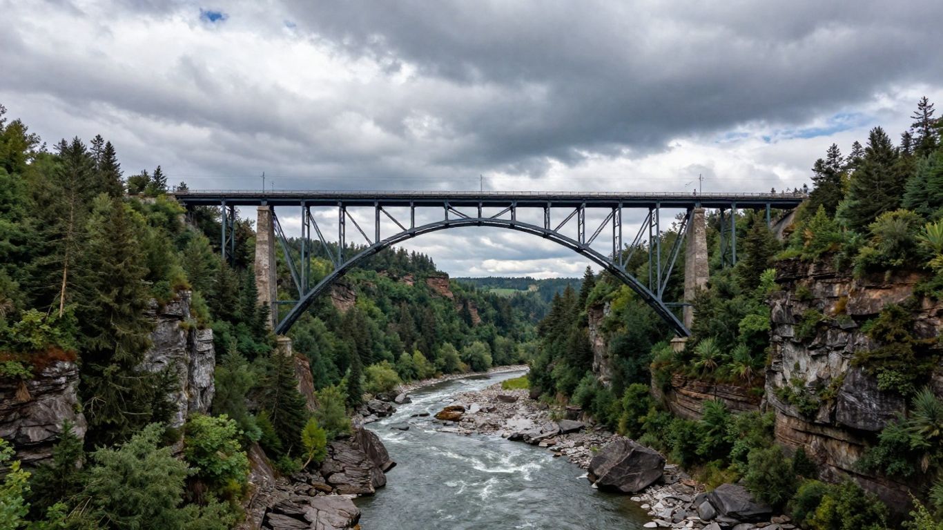 Steel arch bridge spans a rocky river canyon beneath cloudy sky, surrounded by dense evergreen forest