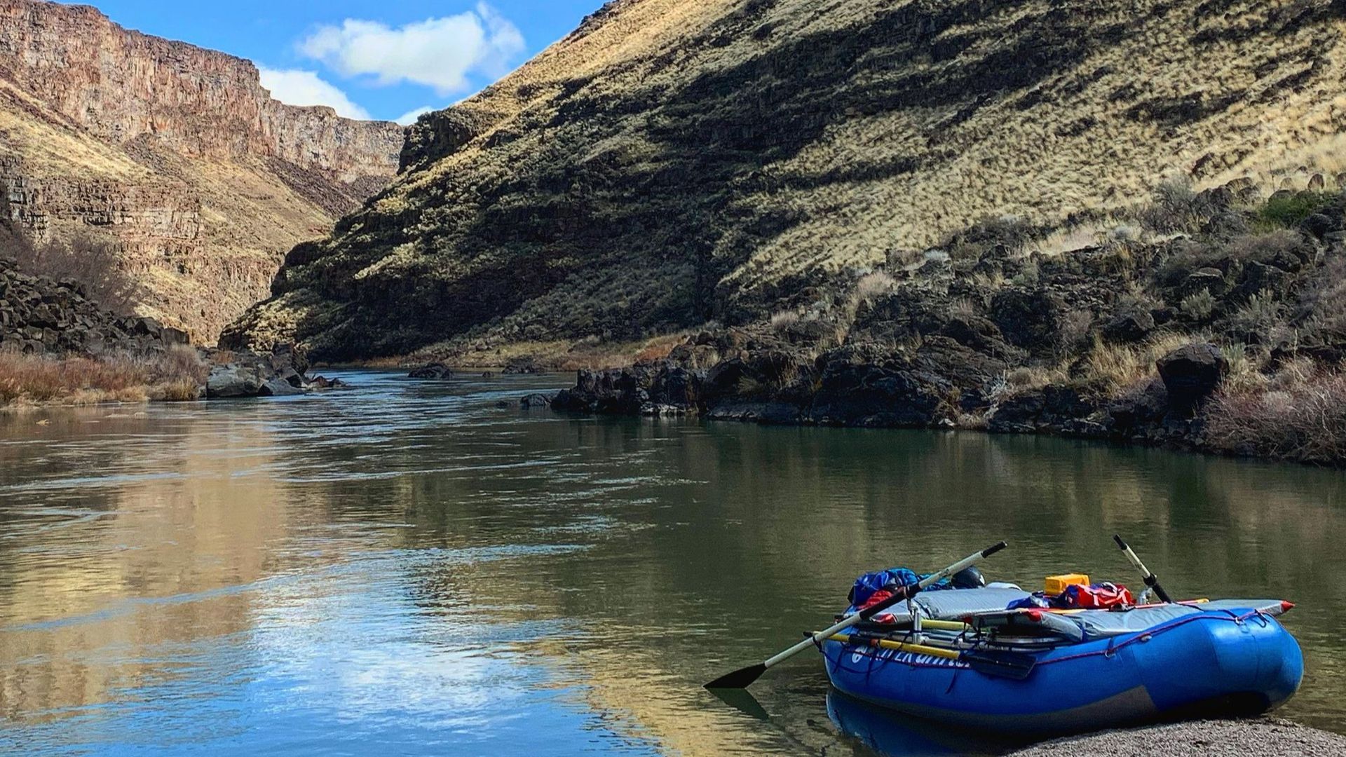 A blue raft floats in a river winding through a sunlit, rocky canyon with dry, grassy hillsides.