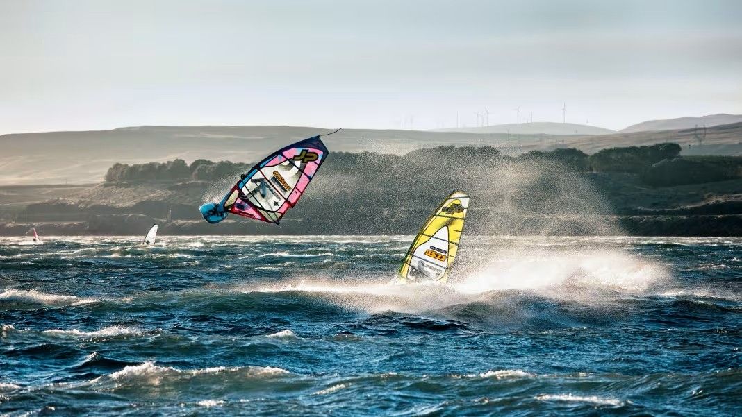 Two windsurfers perform jumps on choppy ocean water under a bright sky with distant hills in the background.