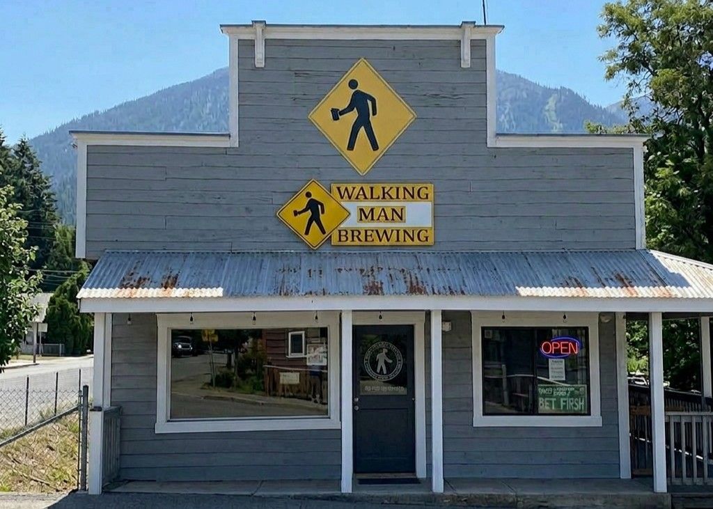 The gray wooden storefront of Walking Man Brewing with yellow pedestrian crossing signs, under a clear blue sky.