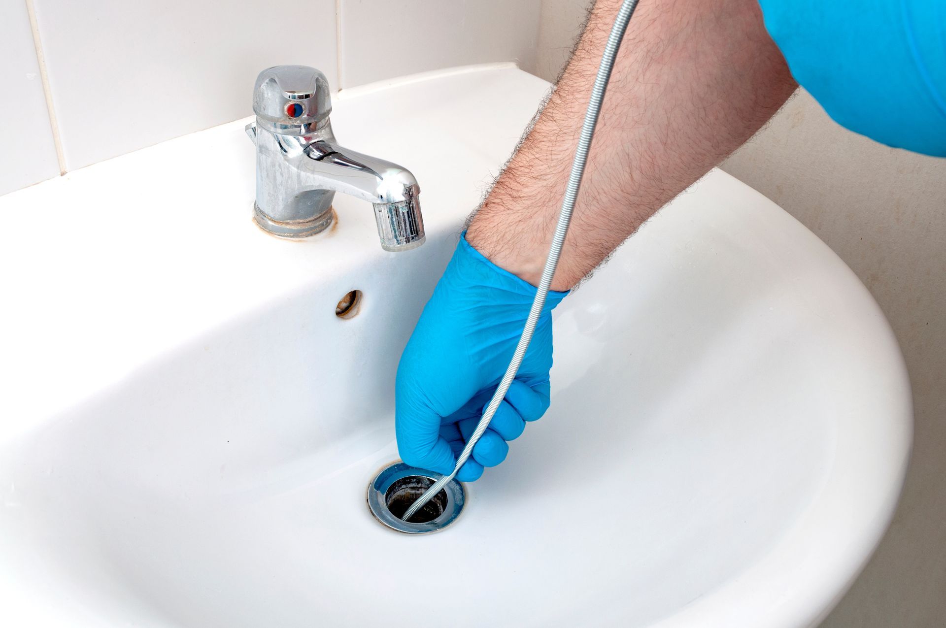 Une personne portant un gant bleu utilise un furet métallique pour déboucher un lavabo blanc.