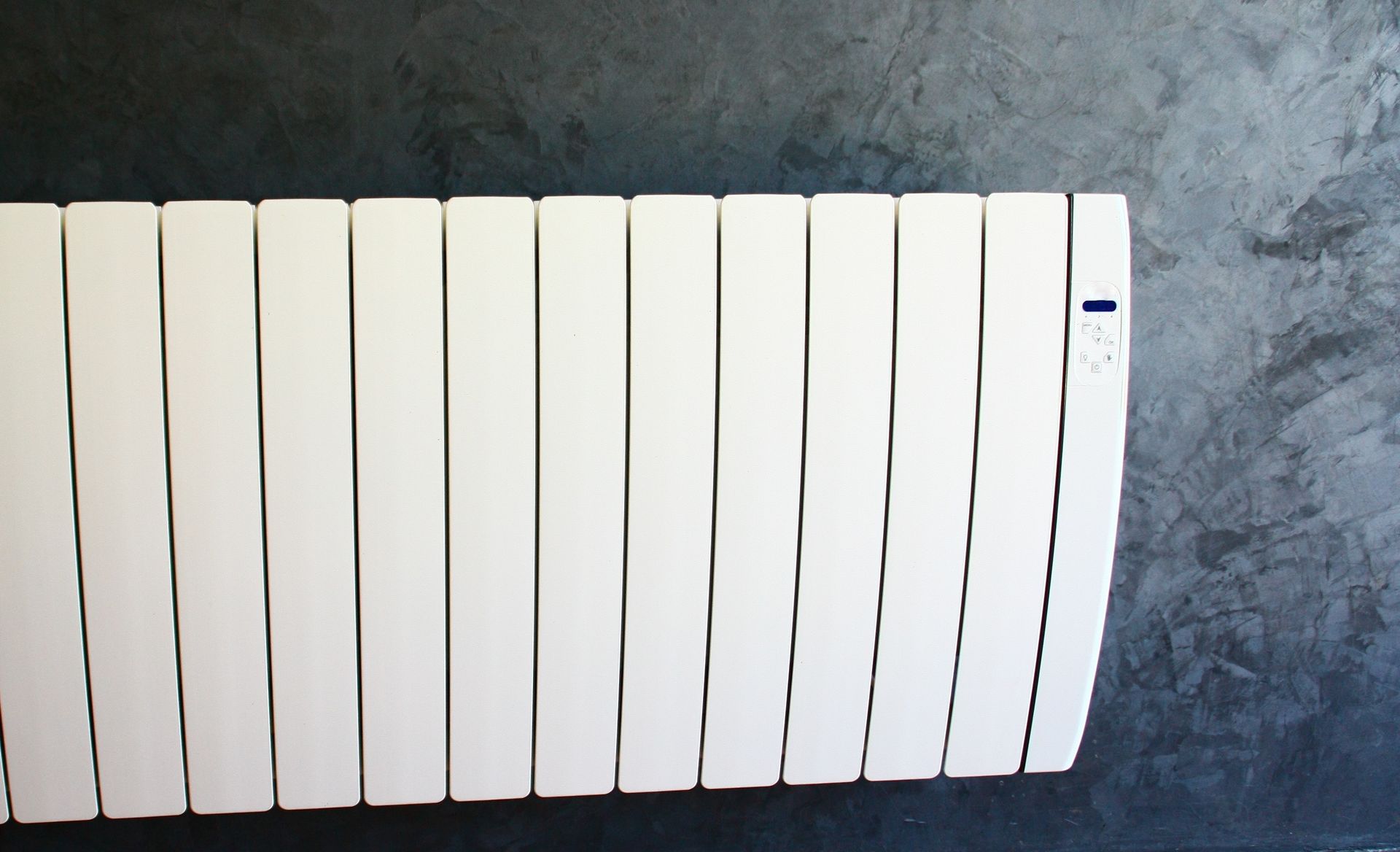 Un radiateur électrique moderne blanc à ailettes verticales, fixé sur un mur texturé gris foncé.