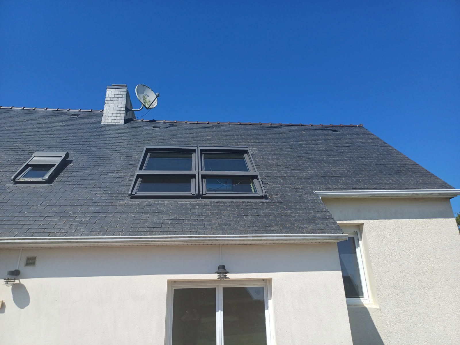 Toit sombre percé de puits de lumière, cheminée et antenne parabolique se détachant sur un ciel bleu azur. Façade blanche du bâtiment en contrebas.