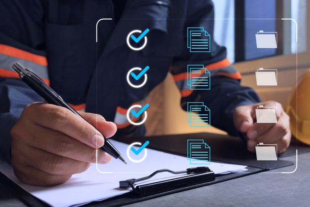 Person in blauer Arbeitskleidung füllt Checkliste mit Häkchen aus, in der Nähe eines Schutzhelms.