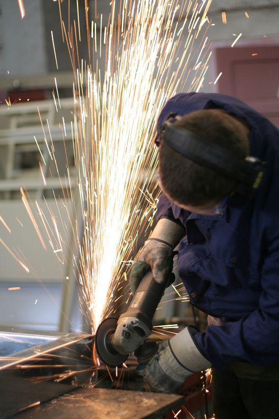 Arbeiter mit Gehörschutz und Handschuhen schleift Metall, während ein starker Funkenflug entsteht.