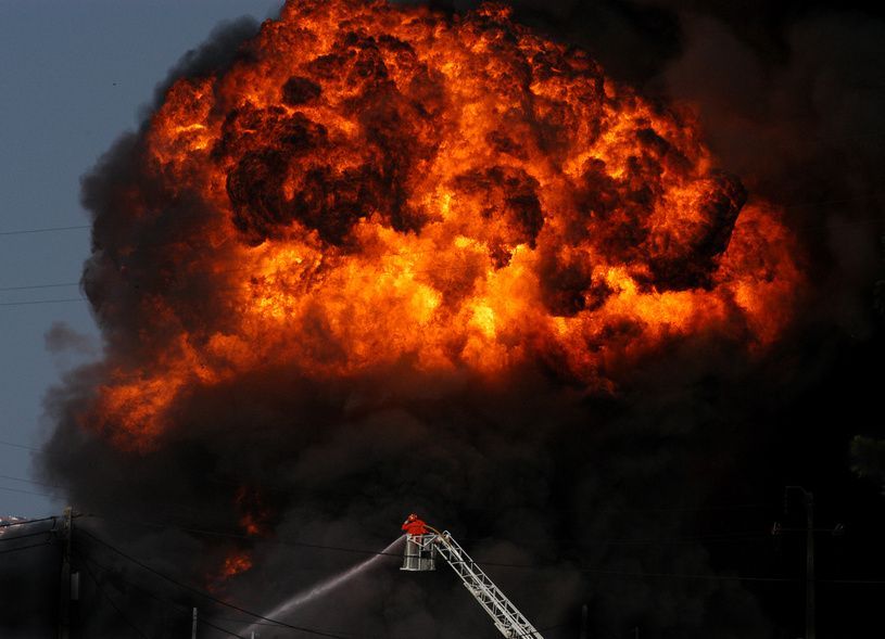 Große Feuerexplosion mit dichter schwarzer Rauchwolke, während ein Feuerwehrmann von einer Drehleiter aus löscht.