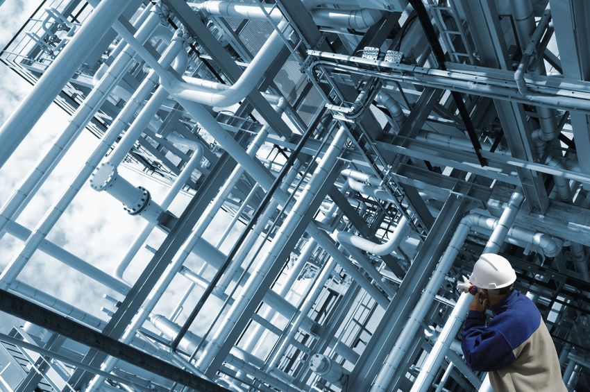 Industrieanlage mit Rohrleitungen und Stahlkonstruktionen, im Vordergrund ein Mitarbeiter mit Schutzhelm beim Telefonieren.