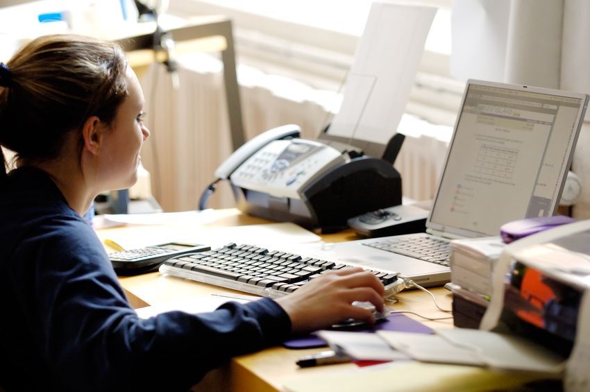 Eine Person in einem blauen Hemd arbeitet an einem Schreibtisch mit Computer, Tastatur und Faxgerät in einem Büro.