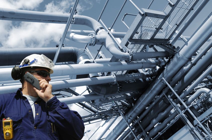 Ein Arbeiter mit Schutzhelm untersucht komplexe Industrierohre und Stahlkonstruktionen in einer blau getönten Raffinerieumgebung.