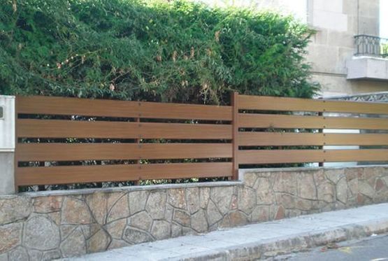 Valla de listones de madera marrón sobre un muro de piedra junto a una acera y vegetación.