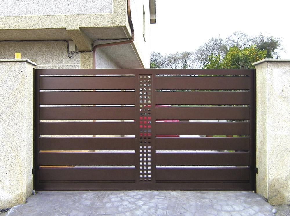 Portón de entrada de metal marrón con listones horizontales entre muros de hormigón y la entrada de la casa.